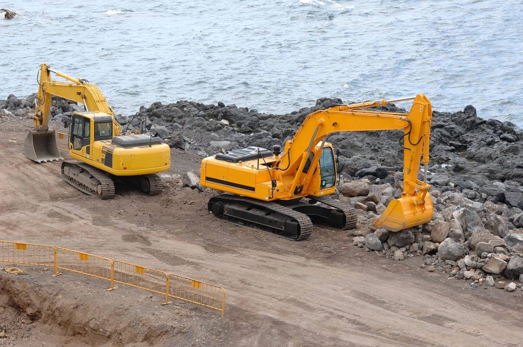 Two excavators on a coastal construction site with rocky terrain.